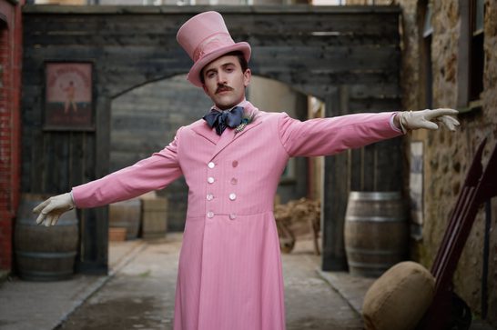 A man in an elaborate Victorian-era costume poses with arms outstretched in a cobblestone courtyard. He wears a bright pink double-breasted frock coat, matching pink top hat, a dark navy bow tie with a peacock feather brooch, and cream-coloured gloves. He has a styled moustache and a confident expression. Wooden barrels and rustic stone and brick buildings are visible in the background.
