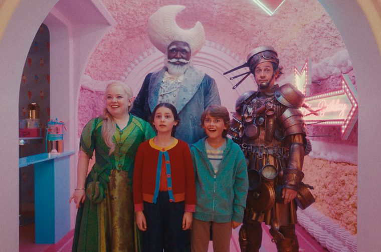 The image shows five characters posed together in a a pink, candy-floss textured environment with arched doorways. A neon sign in the background appears to say something like "Birthday". Left to right, we see a woman in a flowing green dress with long blonde hair, smiling warmly, a young girl in a red/orange cardigan with blue trim, looking up in wonder, a tall man in the centre background wearing a blue coat with a large white crescent moon headdress and a white beard, a young boy in a teal zip-up hoodie, grinning with excitement and a man on the right in elaborate steampunk-style bronze/copper armour, with goggles on his head and various gadgets and pots dangling from his suit.