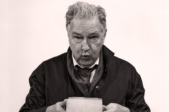 A black and white portrait photograph of an older man, likely in his 60s or 70s, with swept-back grey/silver hair. He has a weathered, expressive face and is leaning slightly forward with an intense, focused expression — his lips slightly parted as if mid-speech or blowing. He's wearing a dark jacket over a shirt and tie, with the tie loosely draped. His hands are visible at the bottom of the frame, holding a small block of ice.