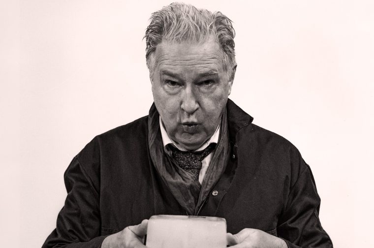 A black and white portrait photograph of an older man, likely in his 60s or 70s, with swept-back grey/silver hair. He has a weathered, expressive face and is leaning slightly forward with an intense, focused expression — his lips slightly parted as if mid-speech or blowing. He's wearing a dark jacket over a shirt and tie, with the tie loosely draped. His hands are visible at the bottom of the frame, holding a small block of ice.