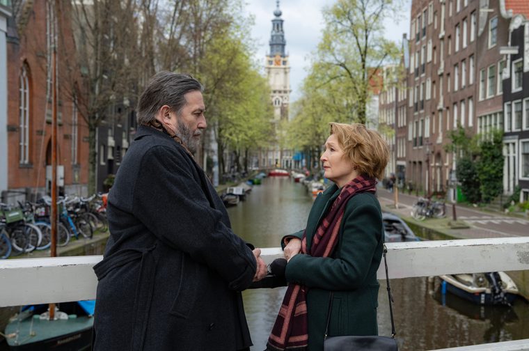 Two middle-aged adults stand facing each other on a bridge over an Amsterdam canal, holding hands. The man wears a dark grey coat and scarf; the woman wears a teal coat with a plaid burgundy scarf. In the background, a historic clock tower rises above tree-lined canal banks flanked by traditional Dutch brick buildings, with boats and bicycles visible along the waterway.