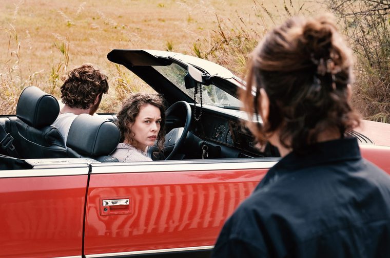 A woman with curly brown hair sits in the driver's seat of a red convertible with the top down, looking over her shoulder with a tense expression. A man with curly hair sits in the passenger seat facing away from the camera. In the foreground, a third person with their hair up is seen from behind, wearing a dark jacket. The car is parked near a field of dry golden grass.