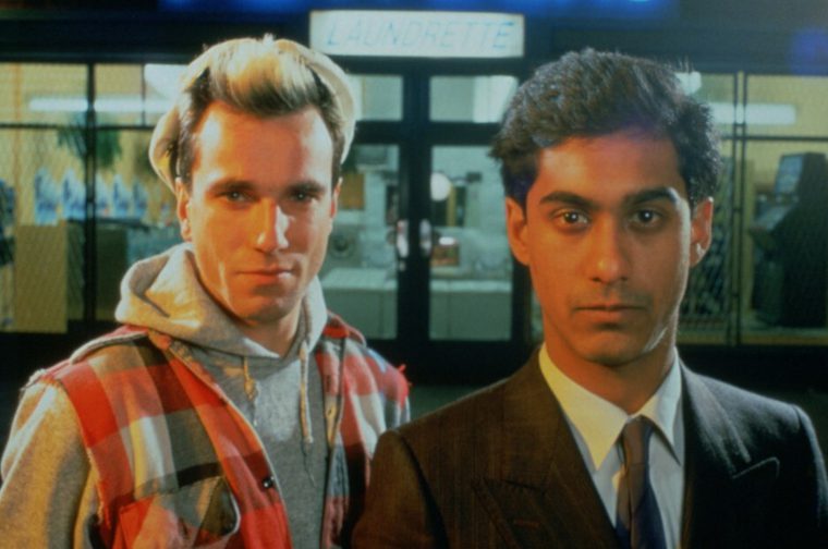Two young men are standing side by side outside a laundrette storefront (the neon "LAUNDRETTE" sign visible above the doorway behind them). The man on the left has bleached blond hair and wears a plaid flannel shirt over a grey hoodie with a necklace visible at the neckline. The man on the right has dark curly hair and wears a formal dark pinstripe suit with a white shirt and striped tie. Both stare directly at the camera with neutral expressions, lit by a warm/blue-toned cinematic glow, with the laundrette's washing machines and fluorescent lighting visible through the glass doors behind them.