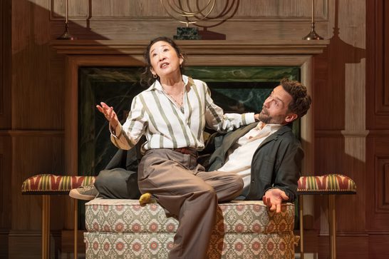 Two actors sit atop upholstered ottomans in front of an ornate wood-paneled fireplace with a green marble surround. An Asian woman with dark curly hair, wearing a green-and-white striped shirt, brown trousers, and layered necklaces, sits cross-legged, gesturing upward with one hand while looking up and speaking animatedly. Beside her, a bearded man in a white shirt and dark green jacket leans back, one arm extended with palm open, looking up in the same direction as if reacting to her words. The setting evokes a wealthy, traditional interior with warm wood tones and gold sconces.