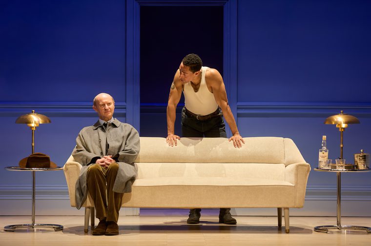 A theatre production still on a minimalist set of saturated cobalt-blue panelled walls, with a dark open doorway at the back. A middle-aged bald man in a grey overcoat, olive corduroy trousers and brown suede shoes sits at the left end of a cream mid-century sofa, hands clasped in his lap, gazing sideways with a wary, composed expression. Behind the sofa, a younger man in a white ribbed vest, dark jeans, boots and a gold chain leans forward on his palms, studying the seated man. Brass mushroom-shaped lamps stand on chrome side tables at either end; a brown fedora rests on the left table, and a bottle of Samovar vodka, two glasses and an ice bucket sit on the right.