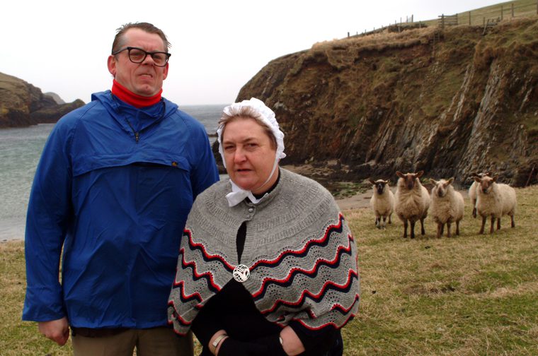 Two people stand on a grassy coastal clifftop on an overcast day. On the left, an older man in a blue windbreaker jacket and red roll-neck top wears glasses. On the right, an older woman wears a traditional grey knitted shawl with red and black zigzag pattern, a white lace cap, and a brooch. Behind them, a small flock of sheep grazes on the grass, with dramatic rocky sea cliffs and a grey, choppy bay in the background.