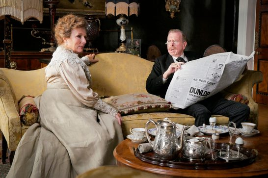 An older man and woman in Edwardian costume are seated on a gold damask sofa in an ornate Victorian drawing room. The woman in a cream lace dress turns to face the man in a dark suit and glasses who is reading a large broadsheet newspaper with the headline 'DUNLOP MANUFACTUR...' visible. A silver tea service is arranged on a round wooden table in the foreground.
