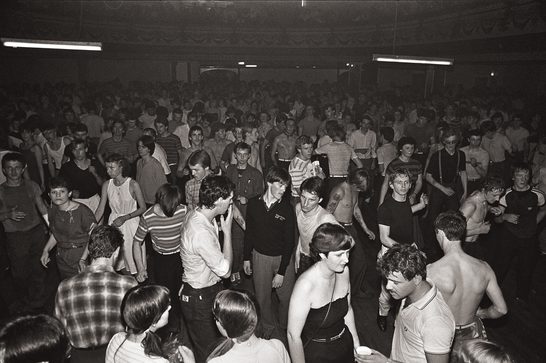 Black and white photograph of a packed dance floor at a Northern Soul all-nighter, showing hundreds of young people dancing and socialising in a large ballroom-style venue, lit by fluorescent strip lights. In the foreground, dancers in casual 1970s clothing — including vest tops, striped shirts, and high-waisted trousers — move energetically, some with shirts removed. The crowd extends deep into the background, filling the hall from wall to wall.