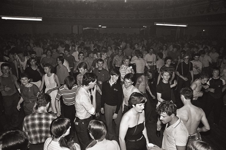 Black and white photograph of a packed dance floor at a Northern Soul all-nighter, showing hundreds of young people dancing and socialising in a large ballroom-style venue, lit by fluorescent strip lights. In the foreground, dancers in casual 1970s clothing — including vest tops, striped shirts, and high-waisted trousers — move energetically, some with shirts removed. The crowd extends deep into the background, filling the hall from wall to wall.