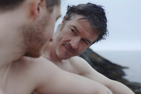 We see two men in an intimate close-up outdoors by the sea. In the foreground, out of focus, one man's bearded jaw and bare shoulder fill the left side of the frame. In sharp focus behind him, an older man with tousled, wet dark hair and stubble looks toward the camera with a faint, knowing smile, his bare shoulders visible. Rocky coastline and a pale, overcast sky and sea are visible in the blurred background.