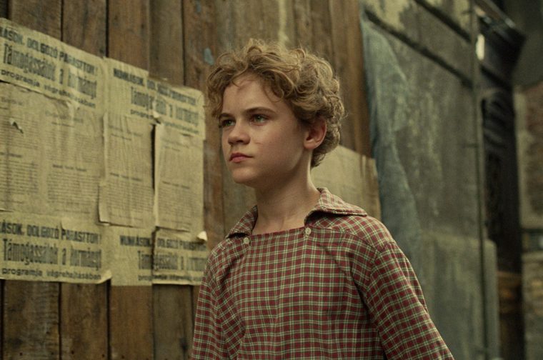 A young boy with curly blonde hair stands by a weathered wooden wall plastered with Hungarian-language posters and notices, wearing a red plaid shirt, with curly blonde hair, gazing pensively to the side in a period drama setting.