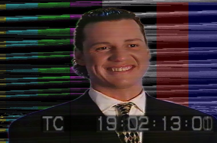 A degraded, analog-video frame showing a man with slicked-back dark hair grinning broadly at the camera. He wears a dark suit jacket, white collared shirt, and a black-and-gold diamond-patterned tie. Behind him, horizontally smeared color bars in yellow, cyan, green, magenta, grey, red, and blue fill the background, with scan-line distortion and tracking noise throughout. A grey on-screen timecode at the bottom reads “TC 19:02:13:00.” The overall look is an eerie, retro broadcast or found-footage aesthetic.
