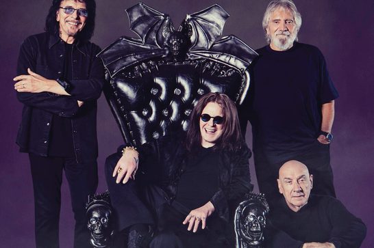 Black Sabbath's four members pose together, with Ozzy Osbourne seated on an ornate black throne featuring bat-wing detailing at its top and skull carvings on its armrests. Osbourne, wearing sunglasses and black clothing, smiles and gestures with his hands. Behind him stand Tony Iommi (with dark curly hair, glasses, and arms crossed) on the left and Geezer Butler (with white hair and beard) on the right. Bill Ward sits to the lower right, bald and dressed in black, resting his chin on his clasped hands. The photo is shot against a dark purple-gray background.