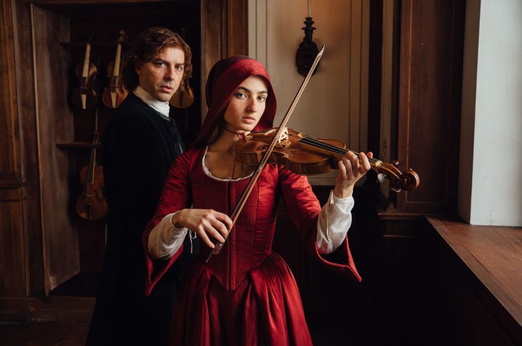 A young woman in a crimson 18th-century dress and matching hood plays a violin, holding the bow with focused intensity, while a man in period black attire stands closely behind her. Several violins hang on a dark wooden wall in the background, suggesting a baroque-era music room or instrument workshop.