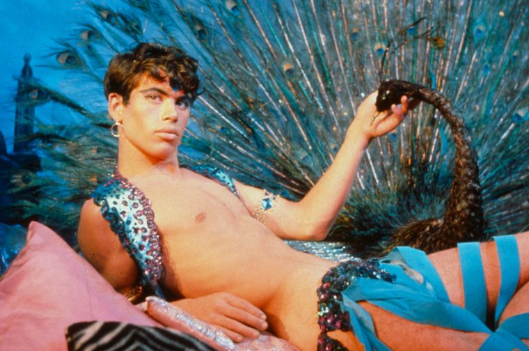 A young white man reclines against pink cushions, shirtless and wearing a jeweled, sequined costume with blue and pink embellishments. He holds a snake aloft in one hand while a large, fan-like peacock feather display spreads behind him in shimmering teal and gold. The scene has a theatrical, fantastical quality with rich, saturated colors.
