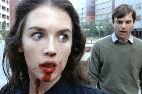 A close-up shot shows a white woman with dark wavy hair and wide, startled blue eyes looking off to the side, with blood smeared across her lips, chin, and neck, dripping onto her dark collar. In the background, slightly out of focus, a man in an olive-green sweater walks toward her with a distressed, open-mouthed expression. The setting is an urban street lined with high-rise apartment buildings under an overcast sky.