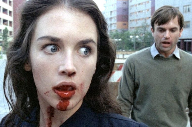 A close-up shot shows a white woman with dark wavy hair and wide, startled blue eyes looking off to the side, with blood smeared across her lips, chin, and neck, dripping onto her dark collar. In the background, slightly out of focus, a man in an olive-green sweater walks toward her with a distressed, open-mouthed expression. The setting is an urban street lined with high-rise apartment buildings under an overcast sky.