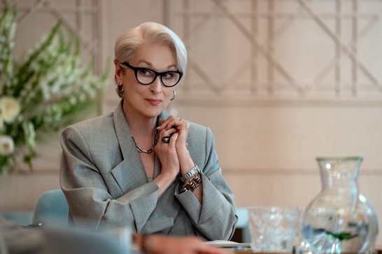 An older woman with short white hair and black cat-eye glasses sits at a table with her hands clasped together in a thoughtful pose. She wears a light grey blazer, a silver chain necklace, hoop earrings, a chunky gold bracelet, and a large ring. The elegant setting includes crystal glassware and a clear glass carafe on the table, with soft white floral arrangements and a panelled wall in the background.