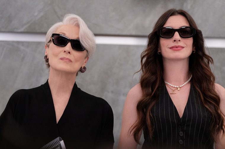 Two women stand side by side against a sleek grey background, both wearing black outfits and large dark sunglasses. The older woman on the left has short silver-white hair and ornate jewelled drop earrings, dressed in a black short-sleeved top and holding a spiral-bound notebook. The woman on the right has long, wavy dark brown hair, wearing a black pinstripe sleeveless waistcoat and layered pearl and gold necklaces.
