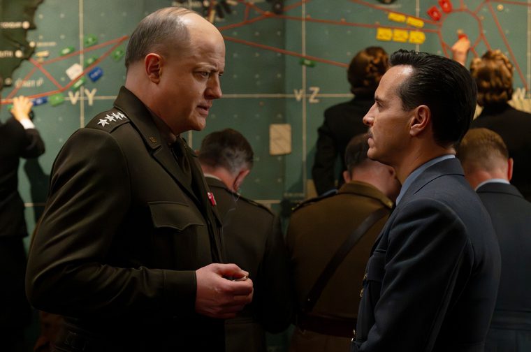 A balding four-star general in a dark green military uniform stands in tense conversation with a middle aged moustached man in a navy suit, both shown in profile facing each other closely. Behind them, a crowded war room is dominated by a large operational map covered in colored markers and route lines, with several other officers and personnel working in the background.