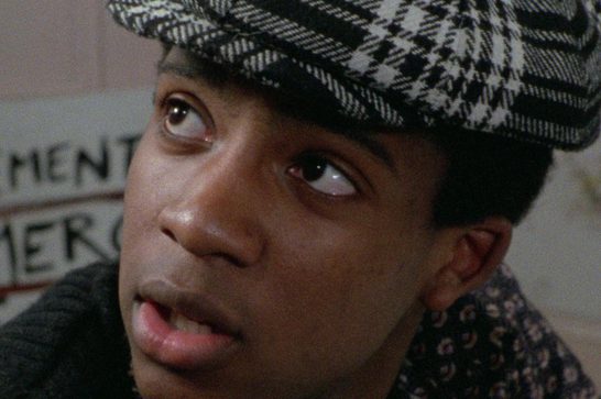 We see a young Black man wearing a black and white houndstooth flat cap, looking upward with a slightly open mouth and a wide-eyed expression. He is dressed in dark and patterned layers. Behind him is a wall with graffiti text partially visible, reading "...GEMENT" and "...MERC..." in large letters.