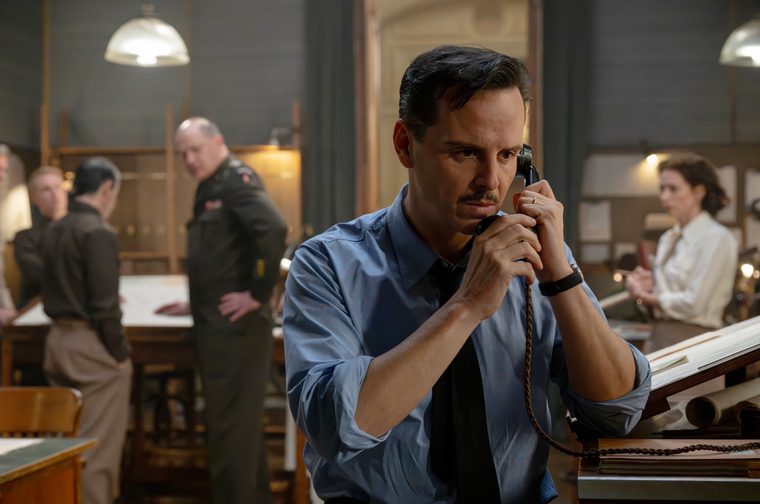 A dark-haired middle aged man with a thin moustache, wearing a light blue shirt with sleeves rolled up and a loosened dark tie, holds an old-fashioned telephone receiver to his ear with a tense, focused expression. He stands in a busy period office or war room filled with drafting tables and blueprints, with a uniformed military officer and several other people working in the background under hanging lamps.
