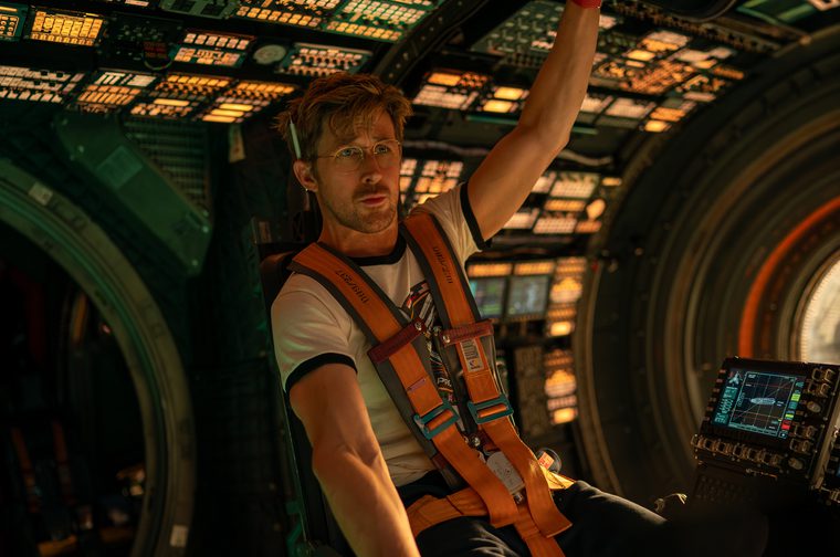 We see a bespectacled man seated in what appears to be a spacecraft cockpit, wearing an orange safety harness over a casual t-shirt and glasses. The cockpit is filled with glowing instrument panels, switches, and screens. His left hand is raised as he holds onto something above him and he also has what looks like a small screwdriver or pen behind his right ear.