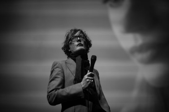 Black and white photo of a man in his fifties performing on stage, holding a microphone with a scarf draped around his neck. He wears thick-framed glasses and a pinstriped jacket, gazing upward with a contemplative expression. His tousled hair falls across his forehead. A large blurred figure or projected image is visible in the background to the right.