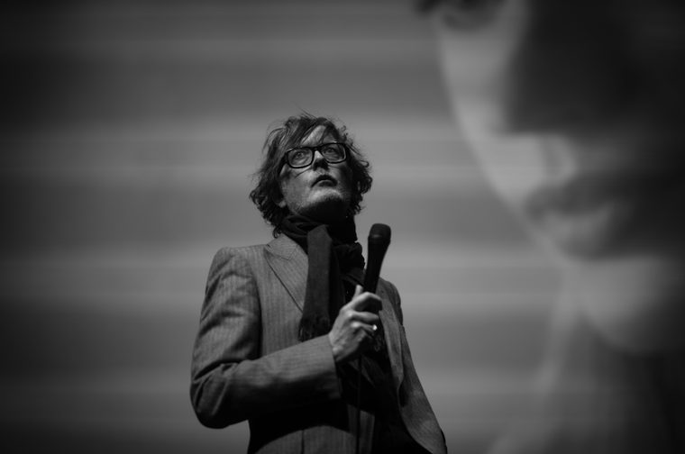 Black and white photo of a man in his fifties performing on stage, holding a microphone with a scarf draped around his neck. He wears thick-framed glasses and a pinstriped jacket, gazing upward with a contemplative expression. His tousled hair falls across his forehead. A large blurred figure or projected image is visible in the background to the right.