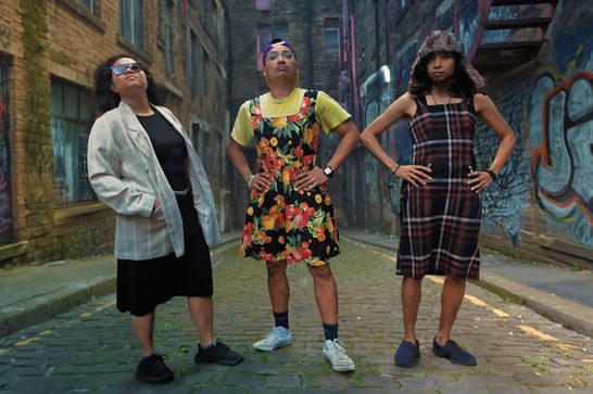 The image shows three individuals of different gender identity posed with hands on hips in a gritty urban alleyway surrounded by brick buildings and colourful graffiti. Their eclectic, gender-nonconforming outfits — a floral pinafore dress over a yellow tee, a plaid dungaree dress with a furry hat, and an oversized blazer with a black skirt — suggest a queer and punk-influenced aesthetic.