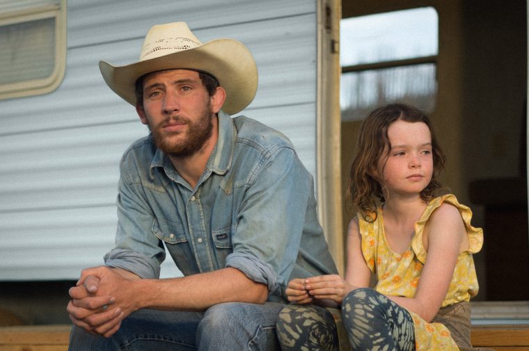 A man in his late twenties with a beard and a cowboy hat on sits down on a step, clasping his hands on his knees. He is wearring a denim short and jeans. A young girl around seven or eight years old sits next to him, wearing a yellow top and blue leggings. Behind them is a mobile home.