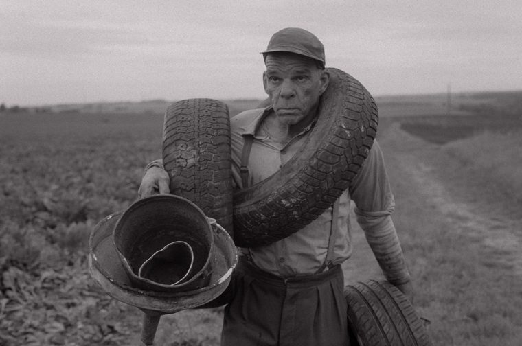 A striking black-and-white film still. An older working man in a flat cap, braces and rolled-up sleeves is hauling multiple worn tyres and a battered metal bucket along a rural dirt track, fields stretching out behind him under a grey sky. He looks directly into the camera with a weathered, stoic expression — burdened but unbowed.