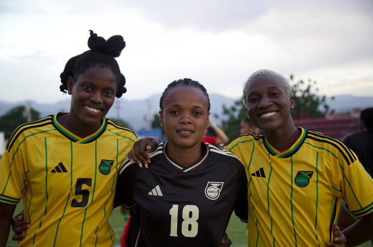 Three young Black women soccer players stand arm in arm on a field, smiling at the camera. Two wear yellow and black striped Jamaica jerseys numbered 5 and an unnumbered shirt, and one wears a black Jamaica jersey numbered 18, with mountains and a cloudy sky in the background.