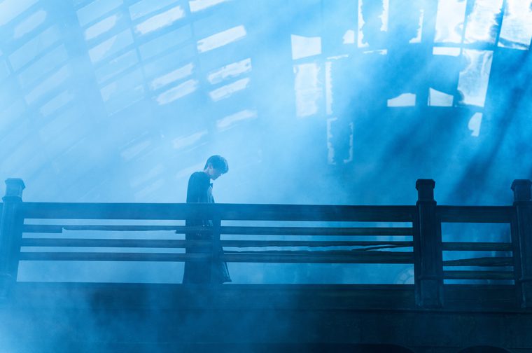 A lone figure stands on a mist-shrouded bridge, bathed in ethereal blue light. Rays of light pierce through what appears to be a large latticed skylight or industrial ceiling above, cutting through thick fog or smoke below. The person — head bowed, dressed in dark clothing — leans against a wooden railing, creating a silhouette that feels deeply contemplative.