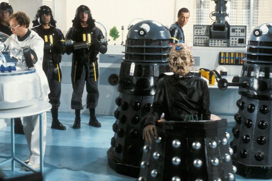 A scarred, cybernetic-eyed figure in black robes sits in a wheeled chair between two black-and-silver Dalek robots, with soldiers in leather uniforms holding gun-staffs and a scientist in a white coat looking on, in a blue-lit laboratory.