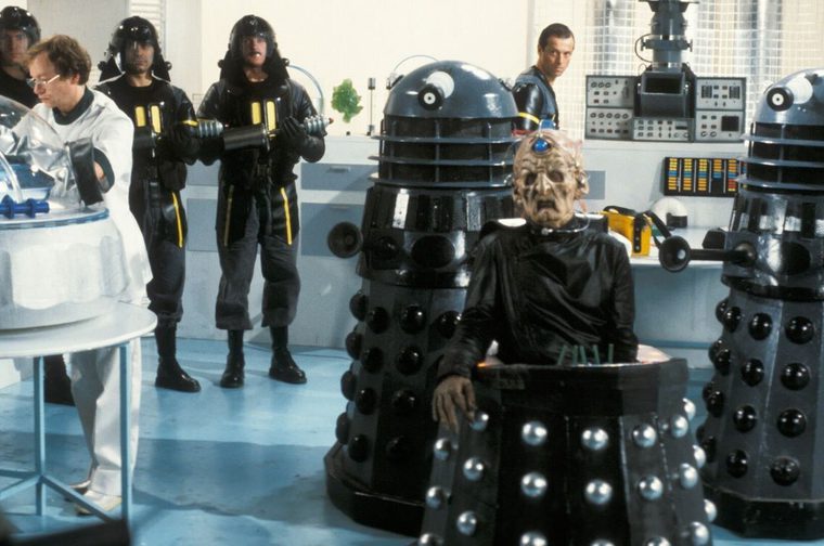 A scarred, cybernetic-eyed figure in black robes sits in a wheeled chair between two black-and-silver Dalek robots, with soldiers in leather uniforms holding gun-staffs and a scientist in a white coat looking on, in a blue-lit laboratory.