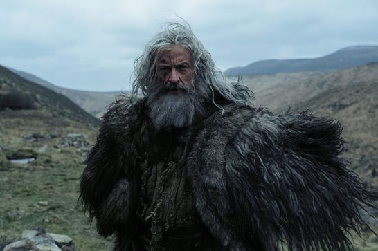 A weathered older man with long, wild grey-white hair and a thick grey beard stands in a windswept, hilly landscape under an overcast sky. His face bears scars and marks, and he wears a heavy, shaggy fur cloak over ragged, layered clothing. Rolling moorland hills fade into misty mountains in the background, with a small stone ruin or cairn visible in the lower left.