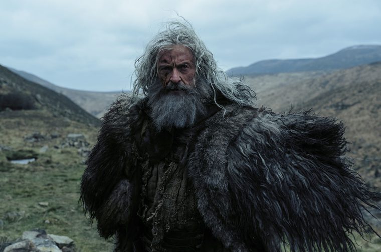A weathered older man with long, wild grey-white hair and a thick grey beard stands in a windswept, hilly landscape under an overcast sky. His face bears scars and marks, and he wears a heavy, shaggy fur cloak over ragged, layered clothing. Rolling moorland hills fade into misty mountains in the background, with a small stone ruin or cairn visible in the lower left.