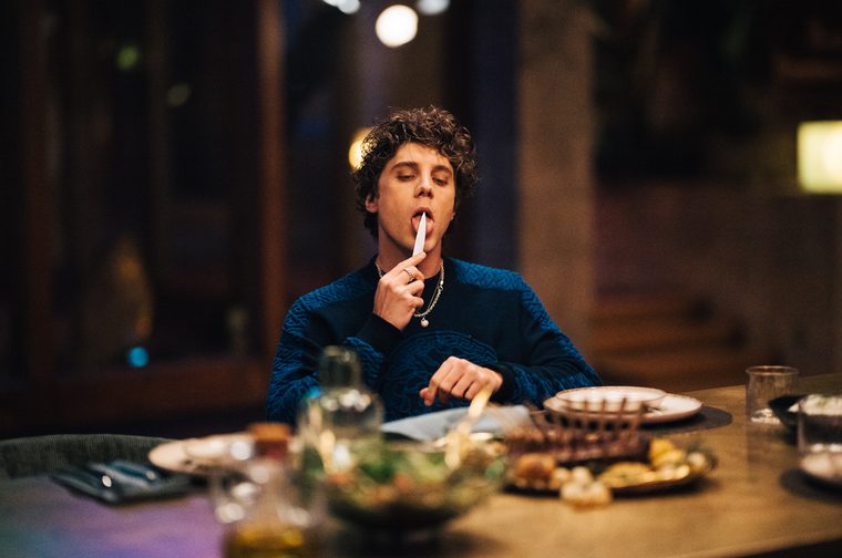 A young person with curly dark hair sits at a dinner table in a dimly lit room, licking a very sharp knife while reaching toward food on the table. They wear a dark blue cable-knit sweater and layered necklaces. The table is set with plates of food, glassware, and a salad bowl, with warm bokeh lights visible in the background.