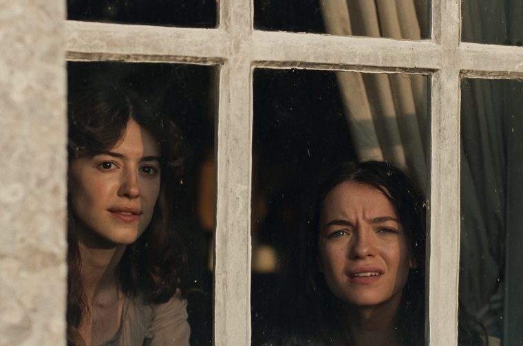 Two young women in period dress peer through the panes of a white-framed window, seen from outside. On the left, a woman with dark wavy hair looks out with a calm, direct gaze. On the right, another woman with dark hair pressed close to the glass has a tense, worried expression, her brow furrowed and mouth slightly open. A curtain is visible behind them in the dim interior.