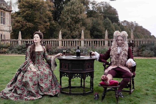 A period drama scene set on the lawn of a grand English country house. Two figures in elaborate 18th-century costumes sit on either side of a dark ornate octagonal table bearing a bottle of red wine and two glasses. On the left, a young woman in a green and red floral silk gown with lace-trimmed sleeves sits on the grass, gazing away from the table. On the right, an older man in a dark red coat, embroidered waistcoat, and a large curled grey periwig sits in a carved wooden chair, his bandaged foot resting on a red cushion — suggesting gout. Behind them, a stone balustrade with obelisk finials borders a formal garden, and the façade of a Jacobean manor house is visible to the left, framed by autumn trees.