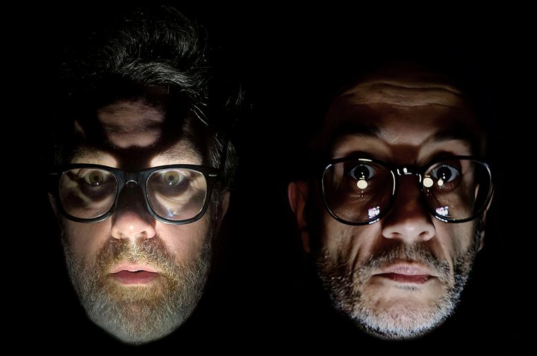 Two disembodied faces float side by side against a solid black background, lit from below in the style of a horror-film portrait. The man on the left has voluminous dark, greying swept-up hair that casts a heavy shadow across his forehead, a full salt-and-pepper beard, and thick square black glasses. His mouth hangs slightly open. The man on the right is bald or close-shaven with a furrowed brow, grey stubble, and round black-rimmed glasses with bright light reflections in the lenses. His lips are pressed tight. Both look directly at the viewer with startled, wide-eyed expressions.