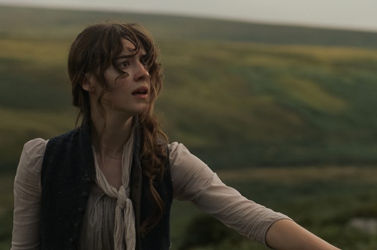 A young woman with wind-tossed, curly reddish-brown hair stands outdoors in period dress — a cream blouse with a tied neck scarf under a dark waistcoat. Her arm is extended outward as she looks off to the side with a startled or urgent expression, her mouth slightly open. Behind her, a soft-focus landscape of rolling green and gold hills stretches under a pale, overcast sky.