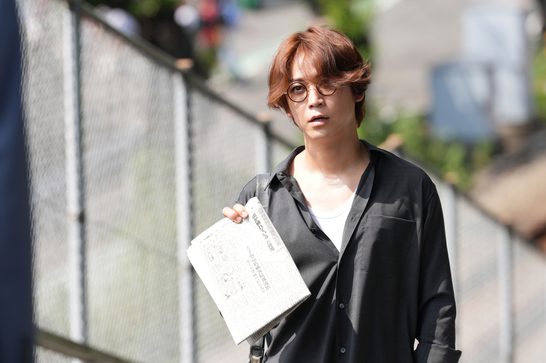 We see a younger Japanese man with auburn/reddish-brown hair and round glasses, wearing a black oversized shirt over a white top, holding what appears to be a Japanese newspaper. The setting looks like an outdoor urban area, with a chain link fence visible behind him.