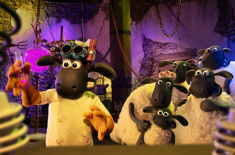 We see a Claymation sheep dressed as a scientist in a white lab coat, wearing goggles and a headlamp pushed up on its head, holding up a glowing pink test tube with hands raised. Behind, a small flock of curious sheep watches, including a smaller lamb peeking out. The background is a spooky, cluttered laboratory or dungeon-like room with stone walls, hanging chains, glowing purple bottles, and eerie purple lighting.