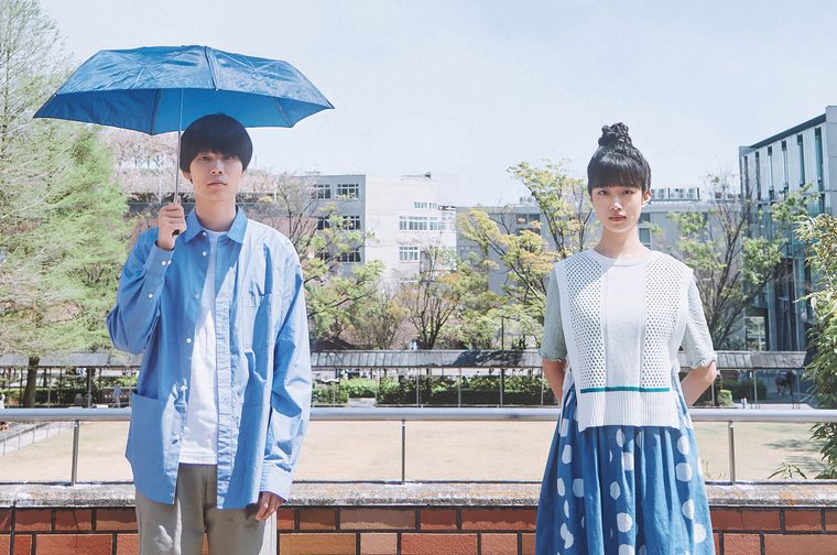 Two young Japanese people stand apart on an outdoor terrace with a metal railing, set against a bright urban campus backdrop with trees and buildings. On the left, a young man in a light blue oversized shirt holds an open blue umbrella despite the clear, sunny sky. On the right, a young woman with her hair in a bun wears a white mesh top and a blue polka-dot midi skirt. Both face the camera with neutral, serious expressions.