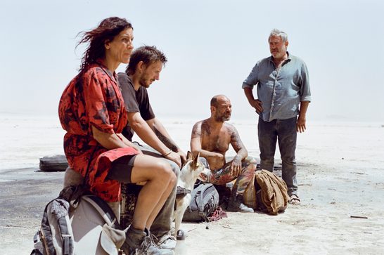 Two men and one woman sit on some luggage in the middle of a barren desert. Another older man stands next to them, his right hand on his hip. The sun beats down relentlessly as one of the sitting men stares at the ground in a forlorn manner.