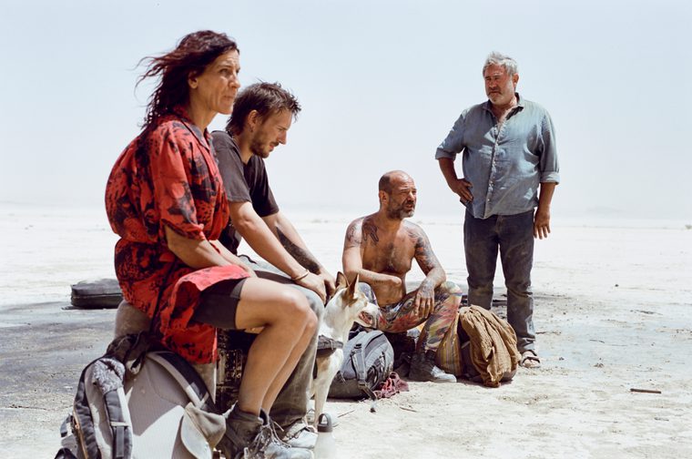 Two men and one woman sit on some luggage in the middle of a barren desert. Another older man stands next to them, his right hand on his hip. The sun beats down relentlessly as one of the sitting men stares at the ground in a forlorn manner.