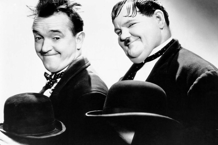 A black-and-white promotional photograph of two men (Laurel and Hardy) in suits and bow ties, each holding a bowler hat. The man on the left has wild, tousled hair and a mischievous grin, while the man on the right is larger, with slicked-back hair, a small moustache, and a broad smile. Both are posed facing the camera in a lighthearted, comedic manner.