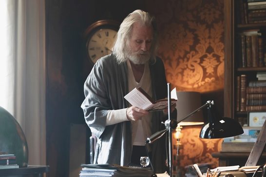 An elderly man with long white hair and a full white beard stands in a book-lined study, reading a small open book he holds in both hands. He wears a white shirt under a gray cardigan and long gray robe. The warm, dimly lit room features a bookshelf packed with leather-bound volumes, an antique wall clock, ornate gold-patterned wallpaper, a black articulated desk lamp, a globe, and stacks of papers and books on a cluttered desk in the foreground.